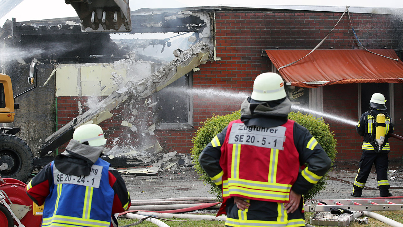 Feuerwehrleute löschen einen Großbrand in einer Halle in einem Gewerbegebiet in Bad Segeberg und reißen dabei mit einem Bagger die Wand eines Hauses ein.