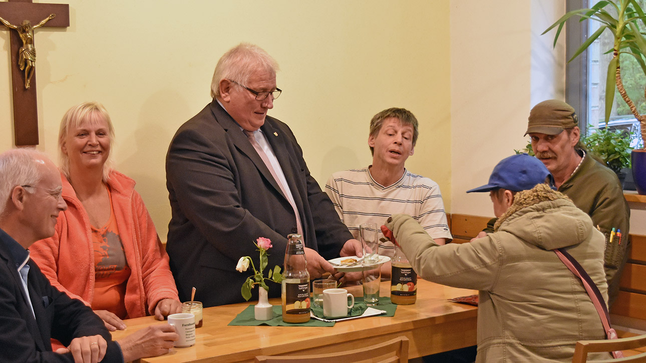 Sechs Personen sitzen an einem Tisch. Landtagspräsident Klaus Schlie reicht bedürftigen Menschen im Speisesaal der Kieler St. Heinrich-Gemeinde einen Teller.