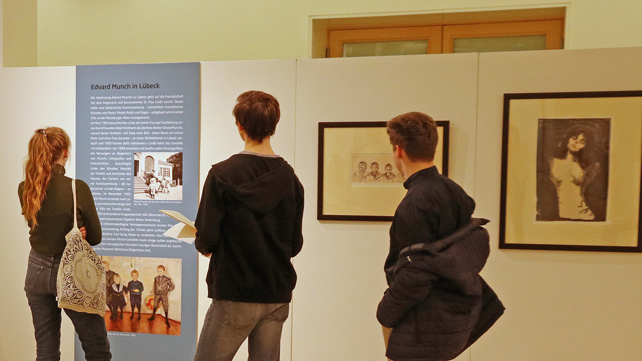 Besucher blicken auf eine Schautafel, die über den Maler Edvard Münch informiert. Daneben sind Edeldrucke von zwei seiner Werke zu sehen.