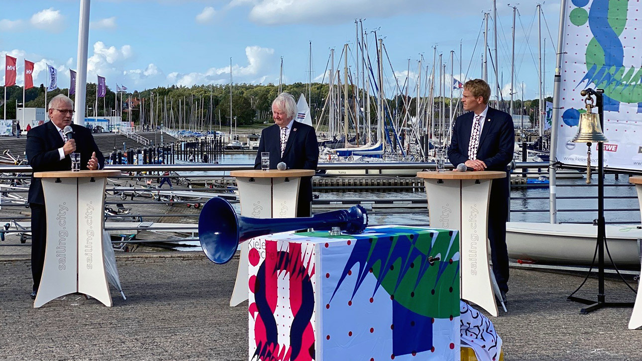 Mit Segelbooten im Hintergrund stehen Landtagspräsident Schlie, Kiels Stadtpräsident Tovar und Ministerpräsident Günther an Stehpulten im Abstand von rund zwei Metern.