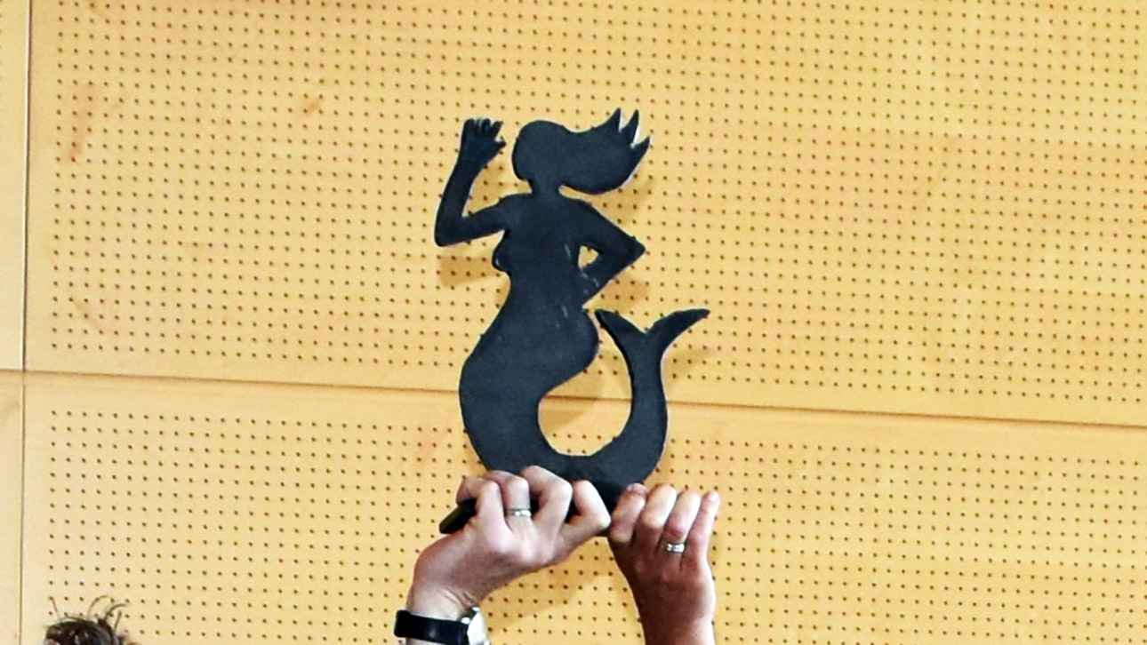 Ein „Emmi för Plattdüütsch in Sleswig-Holsteen“ in Form einer Meerjungfrau wird - getragenen von zwei Händen - in die Luft gestreckt.