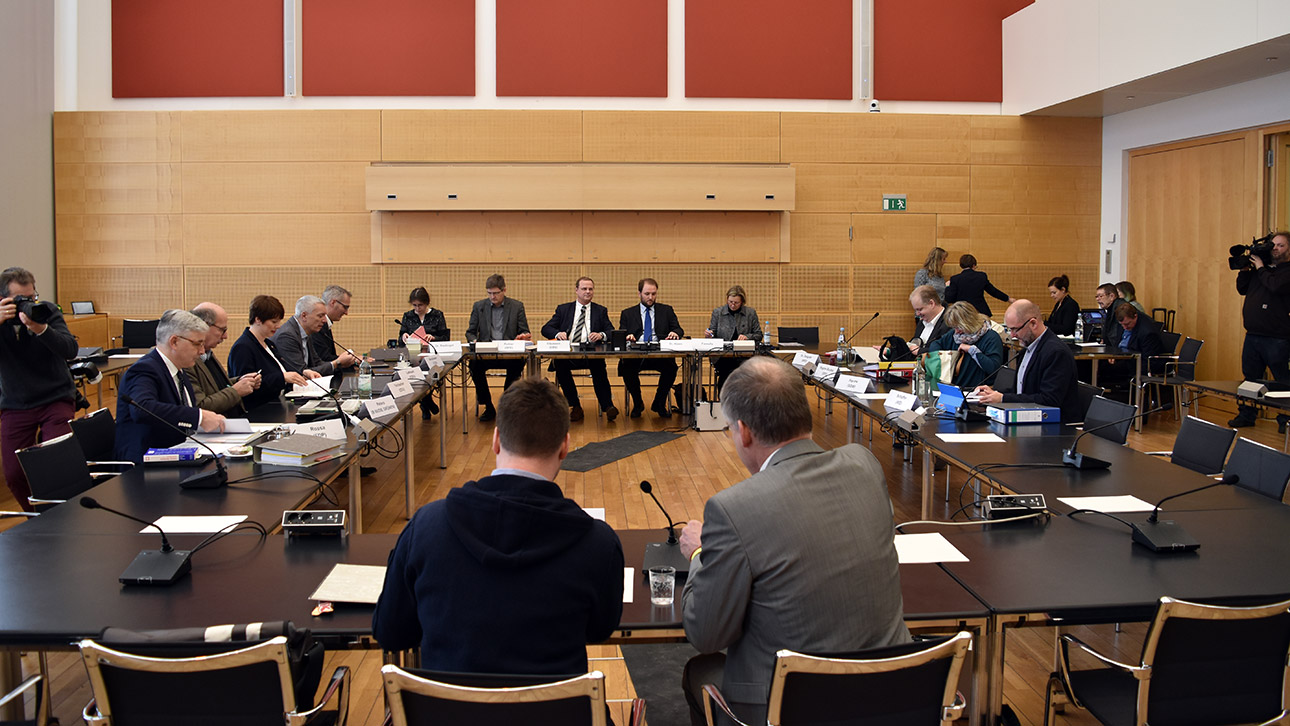 Ein Zeuge mit seinem Rechtsanwalt sitzt mit dem Rücken zur Kamera vor dem Untersuchungsausschuss. Die Abgeordneten sitzen rechts und links an Tischen, der Ausschussvorsitz den Zeugen gegenüber.