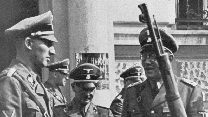 Reinhard Heydrich (links) vor dem Quartier der Einsatzgruppe II in Tschenstochau (Südpolen), Anfang September 1939. neben Heydrich sind zwei weitere uniformierte Personen zu sehen. Das Bild ist in schwrz-weiß.