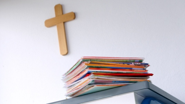 Ein Kreuz hängt in einem Klassenzimmer an der Wand; im Vordergrund liegen Mappen auf einem Tisch.