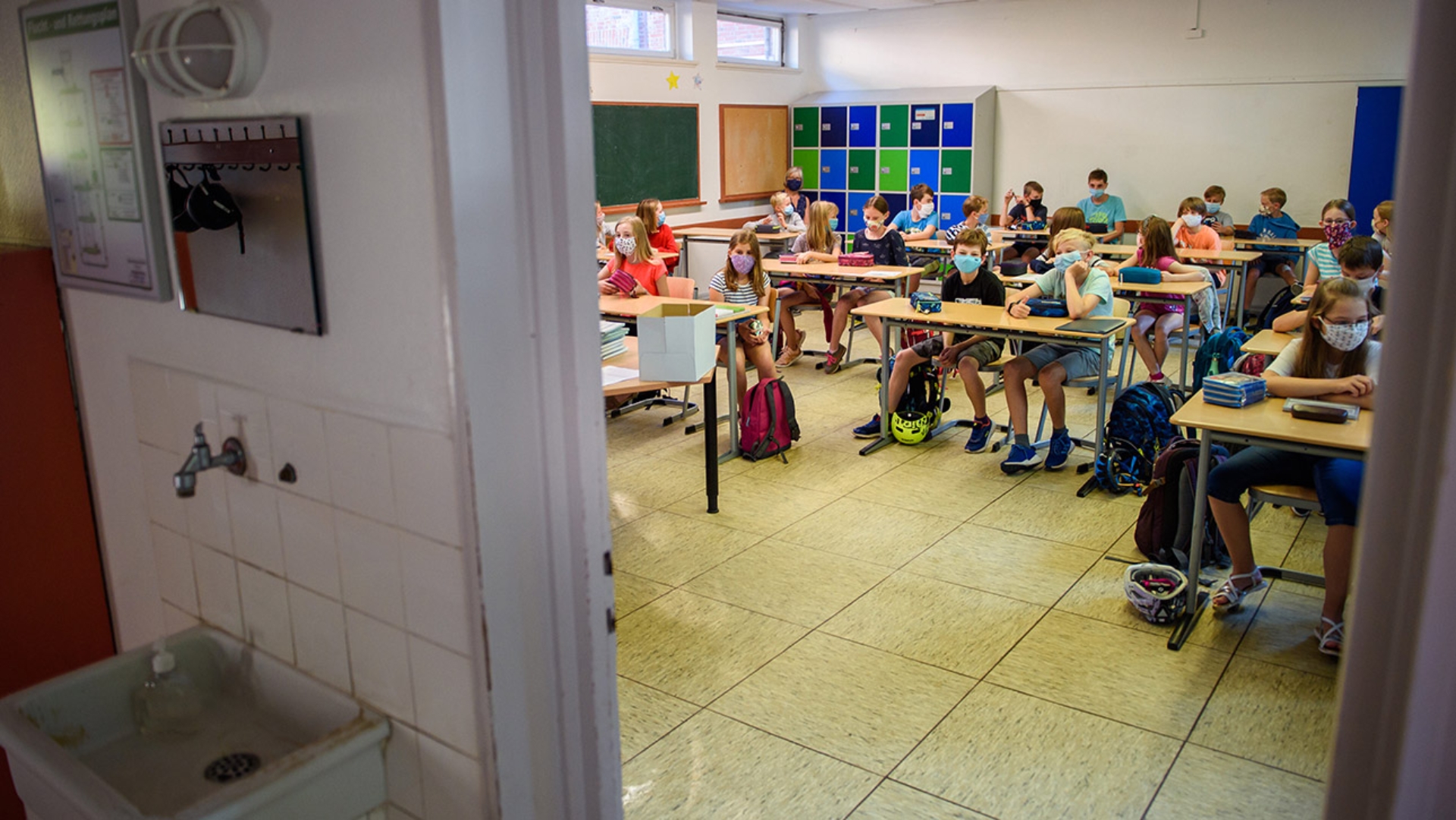 Schüler mit Mund-Nasenschutz einer sechsten Klasse sitzen in einem Klassenraum zusammen. Im Bild ist eine Waschgelegenheit zu sehen.