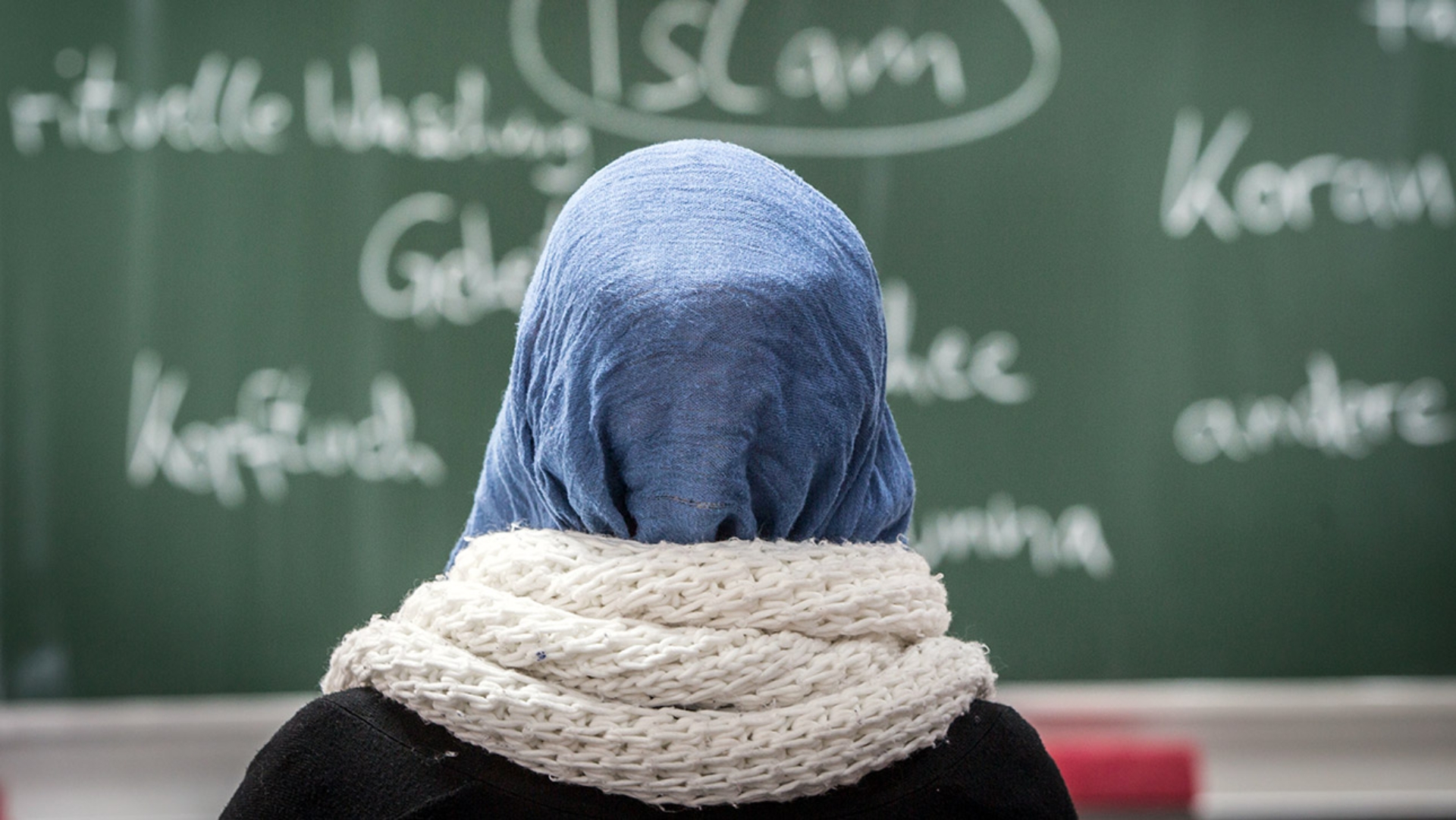 Eine Schülerin mit Kopftuch sitzt in einer Schule bei einer Unterrichtsstunde zum Thema Islam vor einer Tafel.