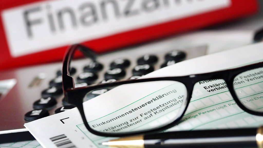 Auf einem Vordruck für die Steuererklärung liegt vor dem Aktenordner mit dem Aufdruck "Finanzamt" ein Stift und eine Brille.