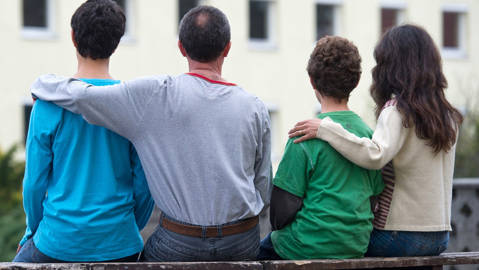 Eine syrische Familie sitzt auf einer Bank vor einem Asylwohnheim einer Zentralen Ausländerbehörde. Mutter, Vater und zwei Kinder haben sich die Arme um die Schultern gelegt.