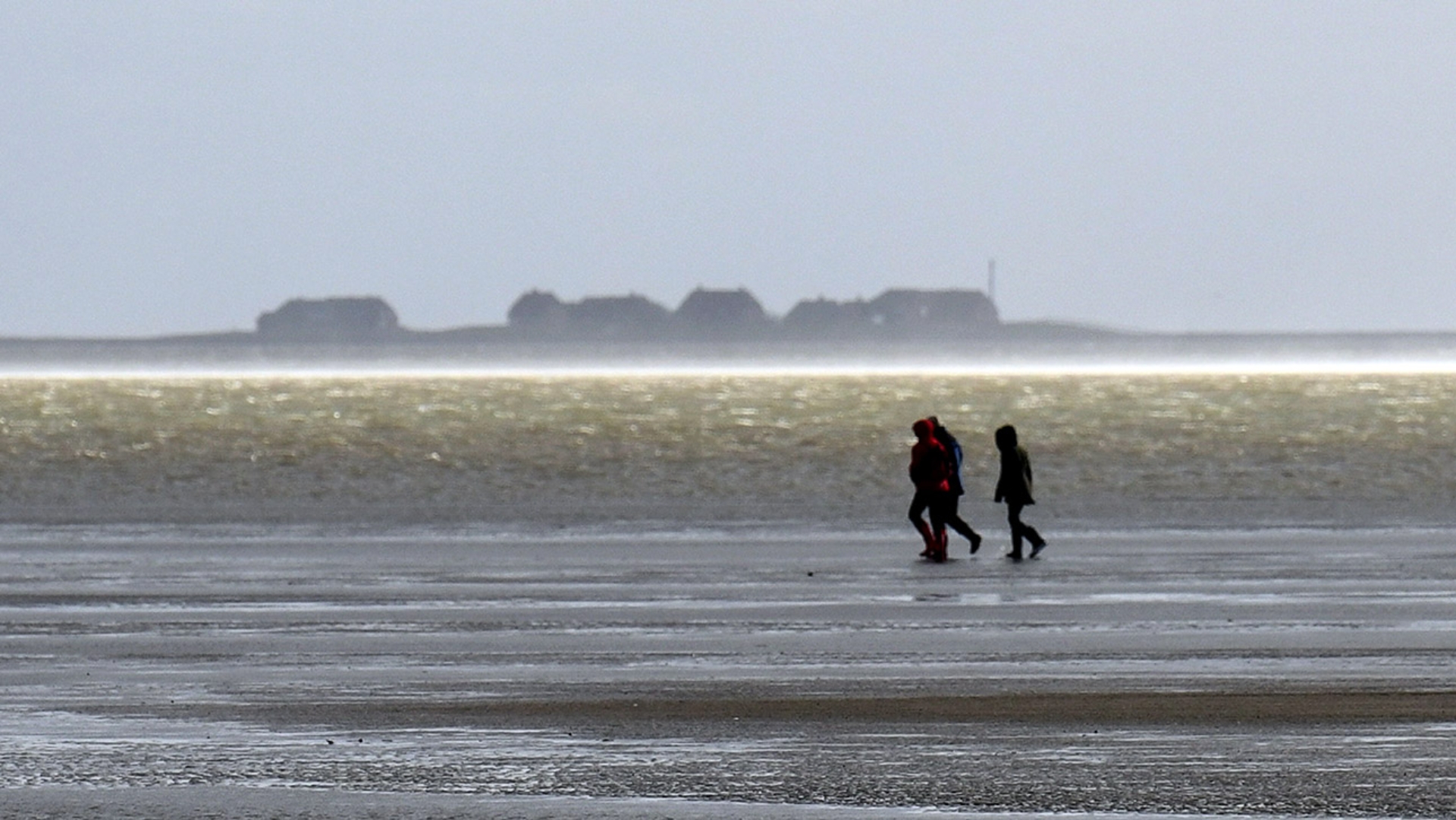 Drei Menschen gehen an der Nordsee spazieren, im Hintergrund Häuser auf einer Insel.