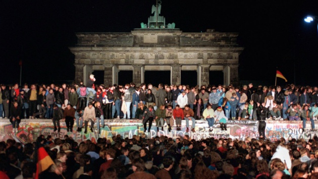 Jubelnde Menschen sitzen auf und stehen entlang der Berliner Mauer am Brandenburger Tor.
