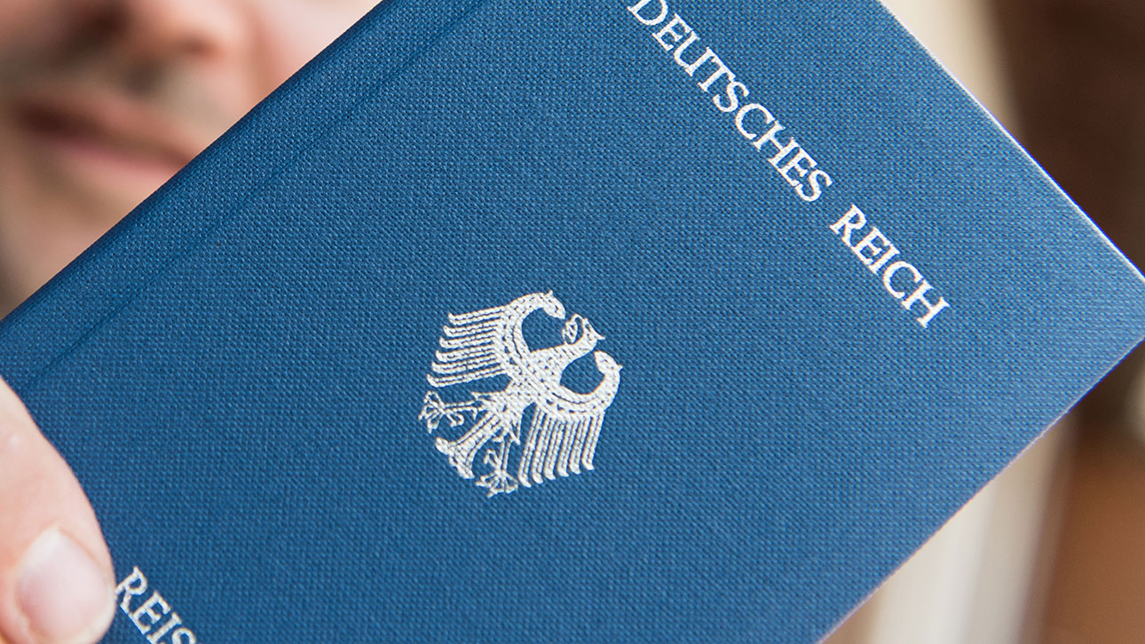 Ein blauer Reisepass wird von einer Hand ins Bild gehalten, auf ihm ist in Goldbuchstaben 