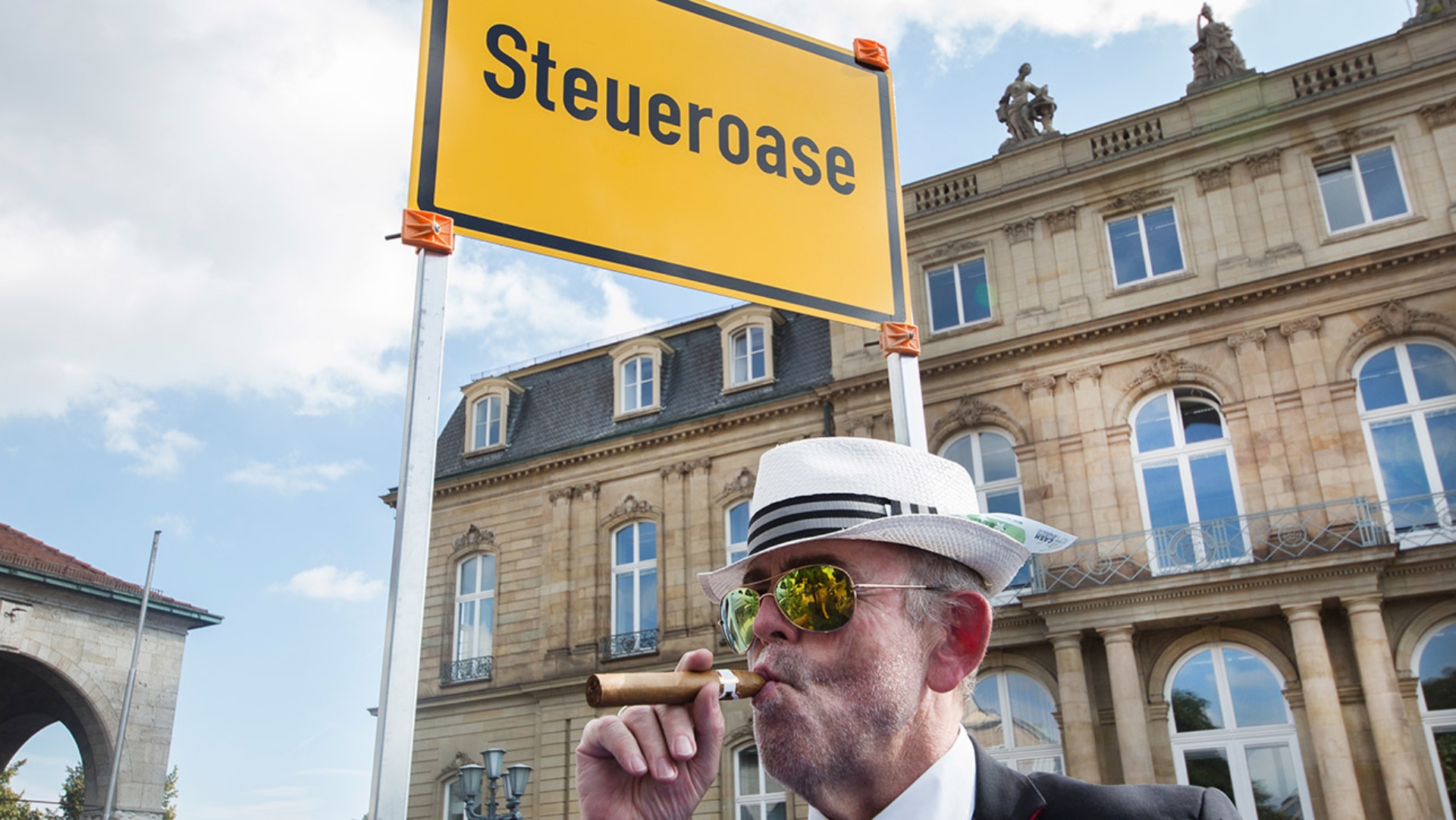 Ein Mann mit Hut, Zigarre und Geldscheinen im Jackett, steht in Stuttgart vor einem Schild  mit der Aufschrift „Steueroase“.