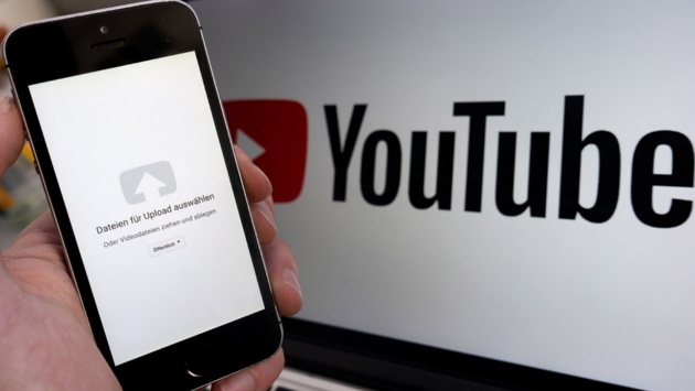 Ein Upload-Fenster der Video-Plattform Youtube ist auf einem Smartphone vor einem YouTube-Logo auf einem Bildschirm zu sehen.