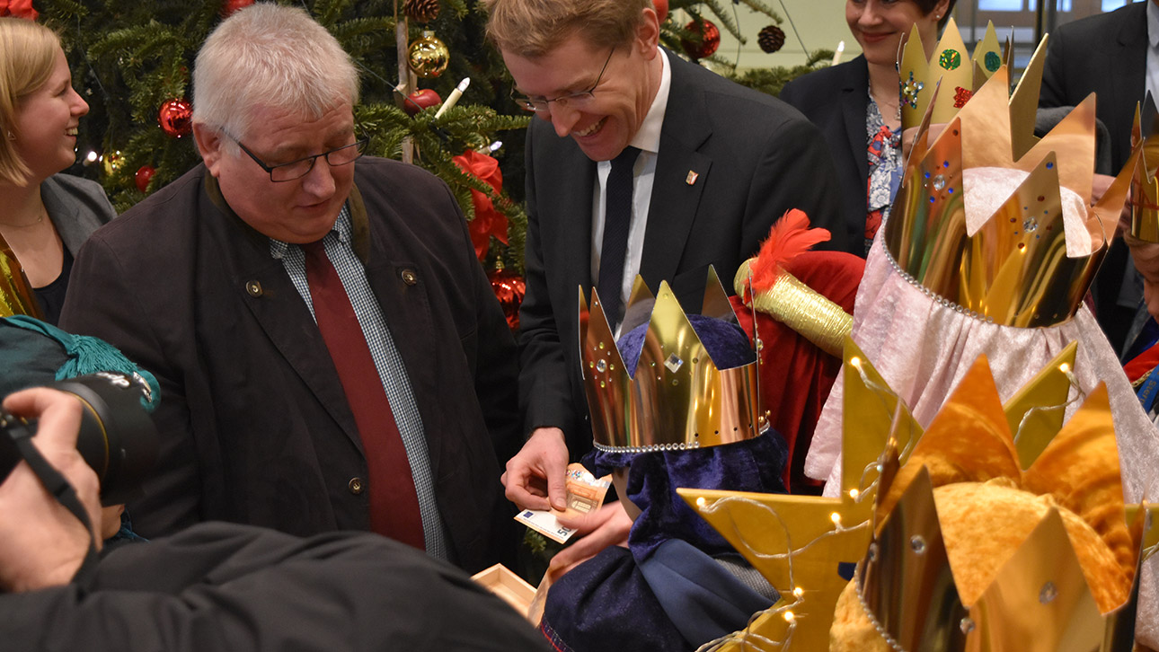 Ein Politiker steckt von Kindern umringt einen 50-Euro-Schein in eine Holzkiste.