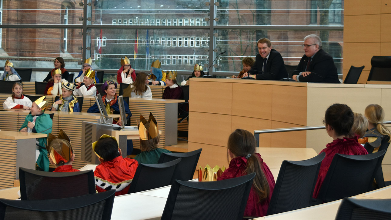 Zwei Politiker sitzen auf den Präsidiumsplätzen im Plenarsaal umringt von Kindern, die als Könige verkleidet sind.