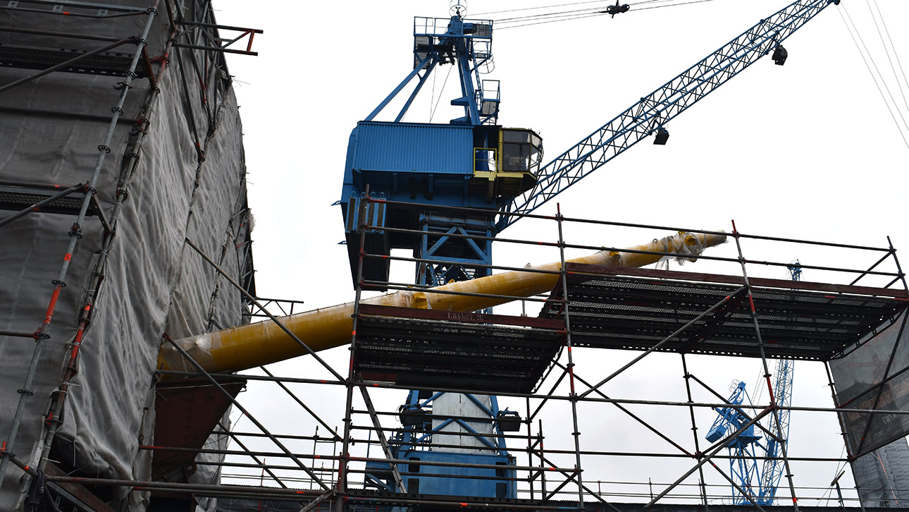 Aus dem Bug des in ein Gerüst gehüllten Segelschiffes ragt eine lange, gelbe Spitze - der Klüverbaum