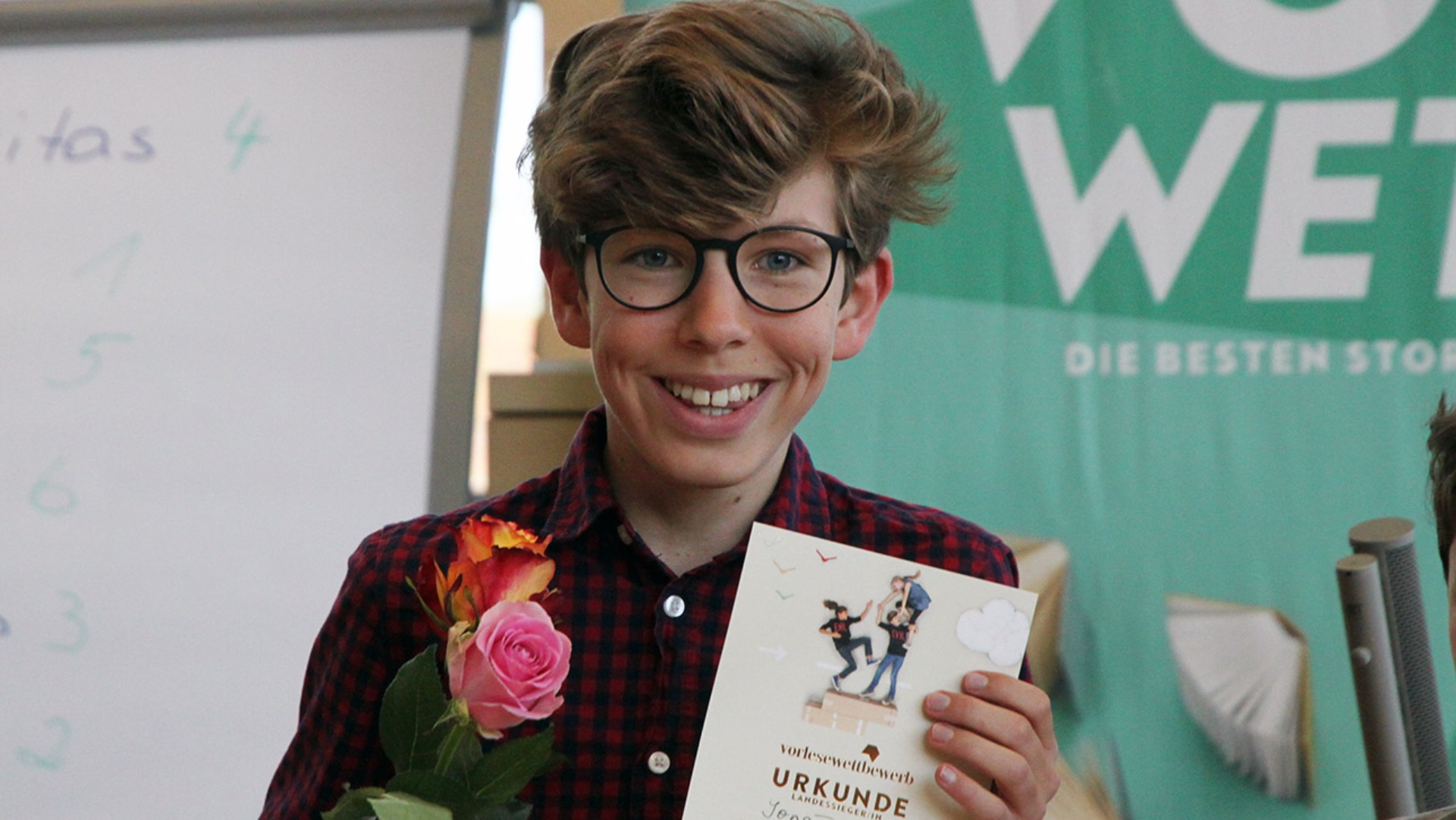 Ein Junge mit einer Rose und einer Urkunde in der Hand lächelt in die Kamera.