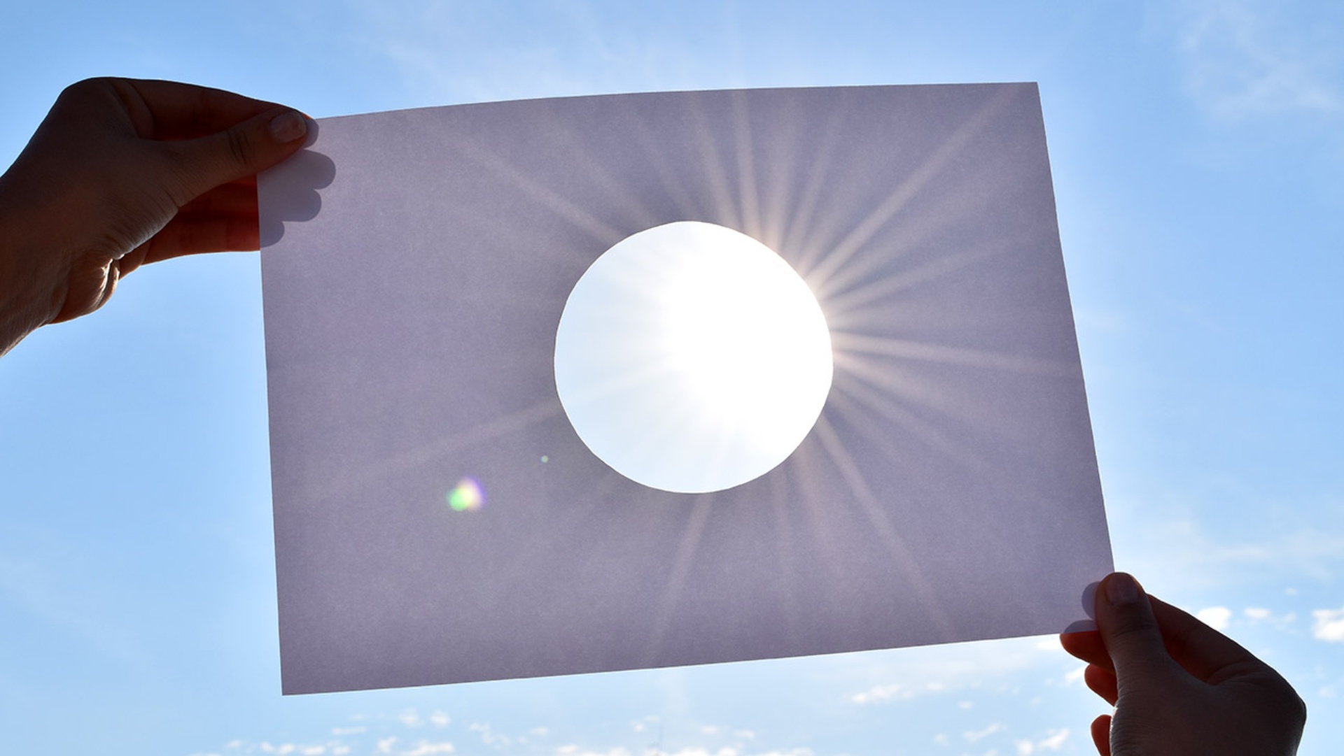 Zwei Hände halten einen Papierbogen mit Loch in der Mitte gegen die Sonne.