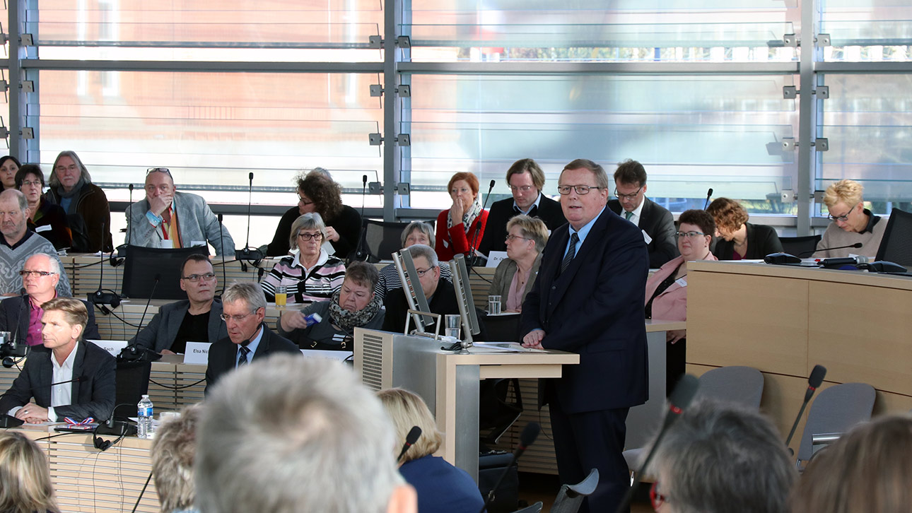 Der Vorsitzende des Sozialausschuss steht am Rednerpult im sonendurchfluteten, vollbestzten Plenarsaal des Landtages