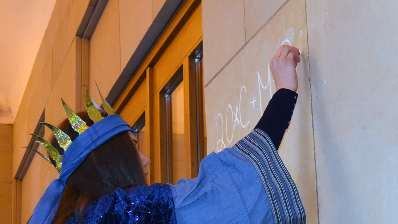 Eine Sternsingerin schreibt mit weißer Kreide „20*C+M+B+19“ an eine Wand am Haupteingang des Landeshauses.