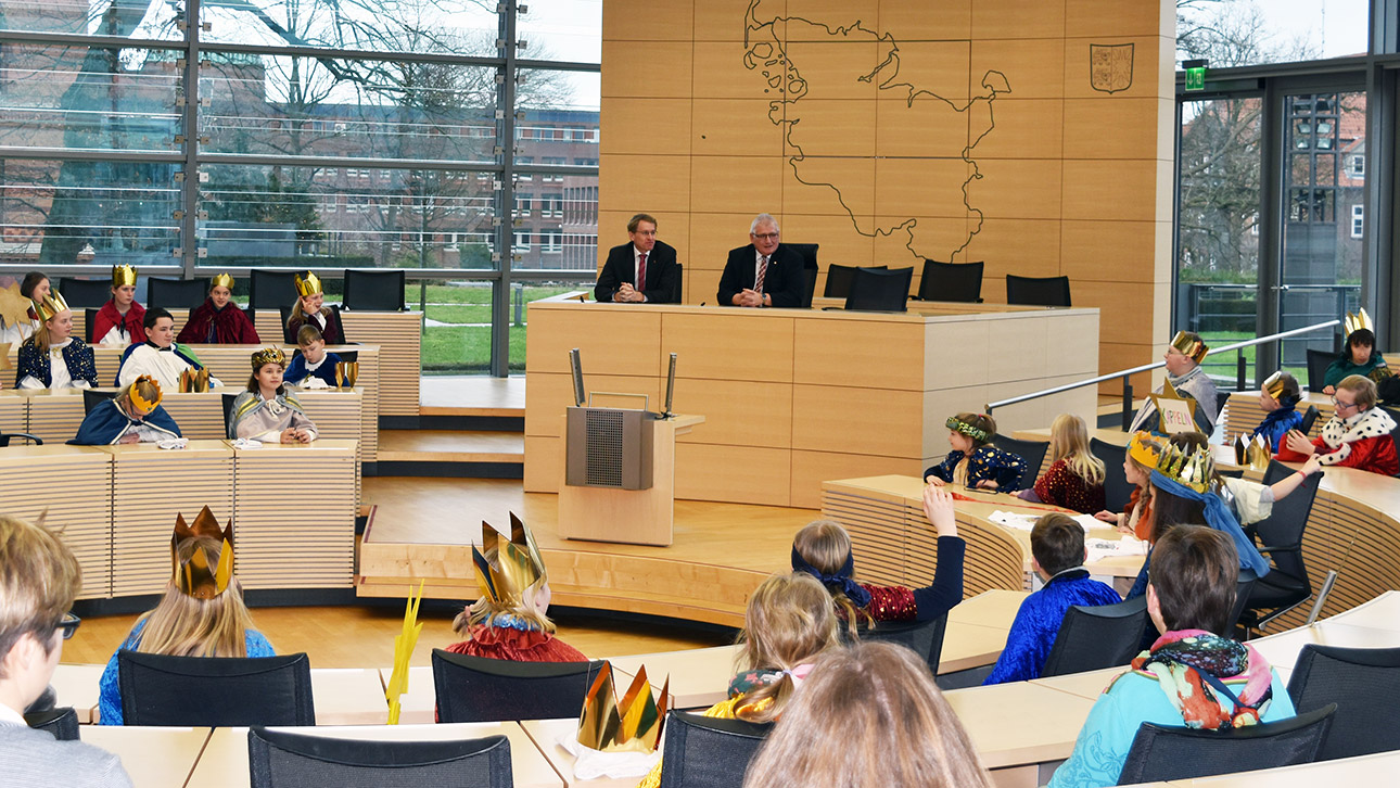 Die Sternsinger sitzen im Plenarsaal. Sie stellen Landtagspräsident Schlie und Ministerpräsident Fragen. Die beiden Politiker sitzen auf dem Präsidialpodium. 