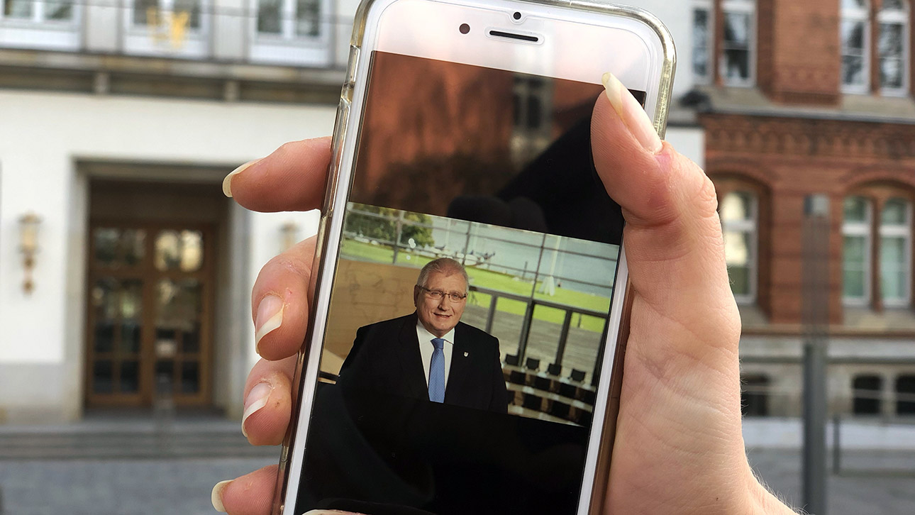 Landtagspräsident Klaus Schlie ist auf einem Smartphone-Bildschirm vor dem Landeshaus zu sehen