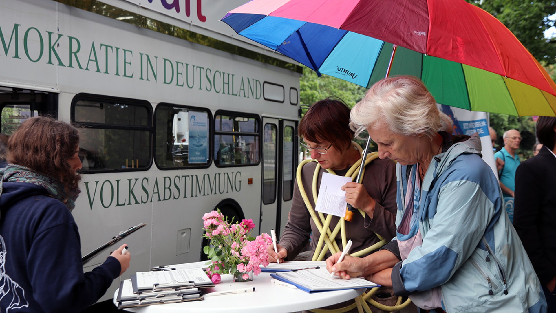 Zwei Passanten tragen sich an einem Stehtisch auf Listen der Volksinitiative eib. Im Hintergund steht ein Doppeldeckerbus, der für die Volksinitiative wirbt.