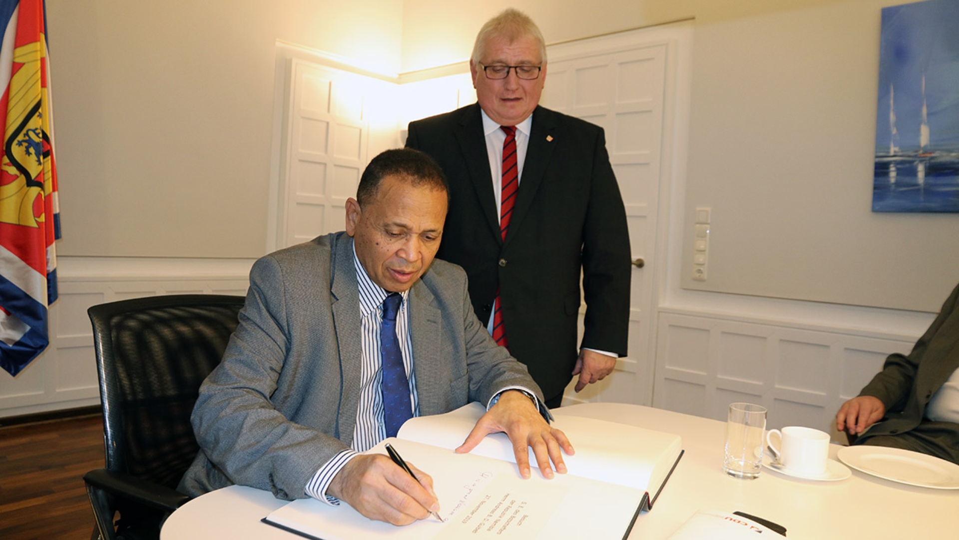 Der Botschafter der Republik Namibia, Andreas B. D. Guibeb, trägt sich in das Gästebuch des Landtages ein. Landtagspräsident Klaus Schlie schaut stehend zu.