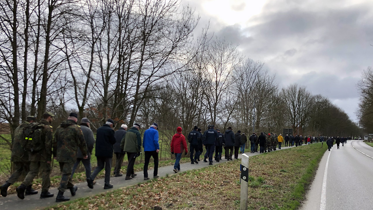 Teilnehmer des Oeversee-Gedenkmarsches gehen in einer langen Schlange an einer Bundesstraße entlang.
