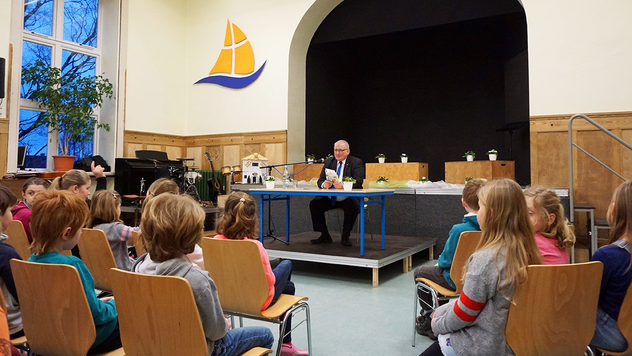 Landtagspräsident Klaus Schlie sitzt auf einem kleinen Podium in der Aula der Christlichen Schule und liest rund 70 Grundschülern aus einem Kinderkrimi vor.