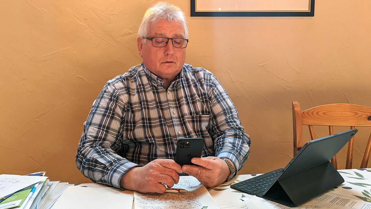 Landtagspräsident Klaus Schlie sitzt an seinem Esstisch im Homeoffice mit viel Papier vor sich und dem Smartphone in der Hand.
