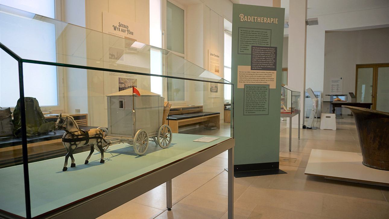 Blick in die Ausstellung zum Thema Seebäderkultur. Zu sehen ist ein Glaskasten mit einem Modell eines sogenannten Badekarren, einer Art fahrbarer Umkleidekabine
