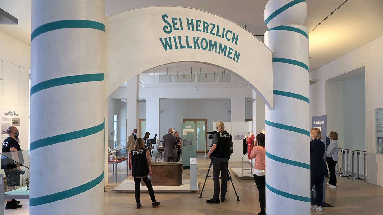 Hinter einem Torbogen in blau-weiß mit der Aufschrift "Sei herzlich willkommen" stehen Besucher in einer Ausstellung zum Thema Seebäder in Schleswig-Holstein.