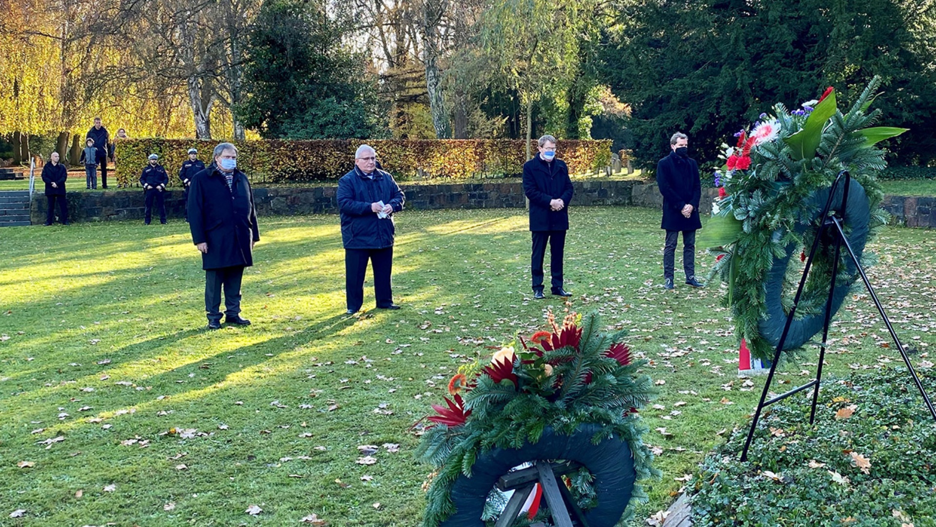 Ekkehard Klug von der Kriegsgräberfürsorge, Landtagspräsident Schlie , Ministerpräsident Günther und Kiels Oberbürgermeister Klaus Kämpfer stehen auf einer Wiese vor mehreren Kränzen und gedenken der Opfer von Kriegen und Gewaltherrschaft.