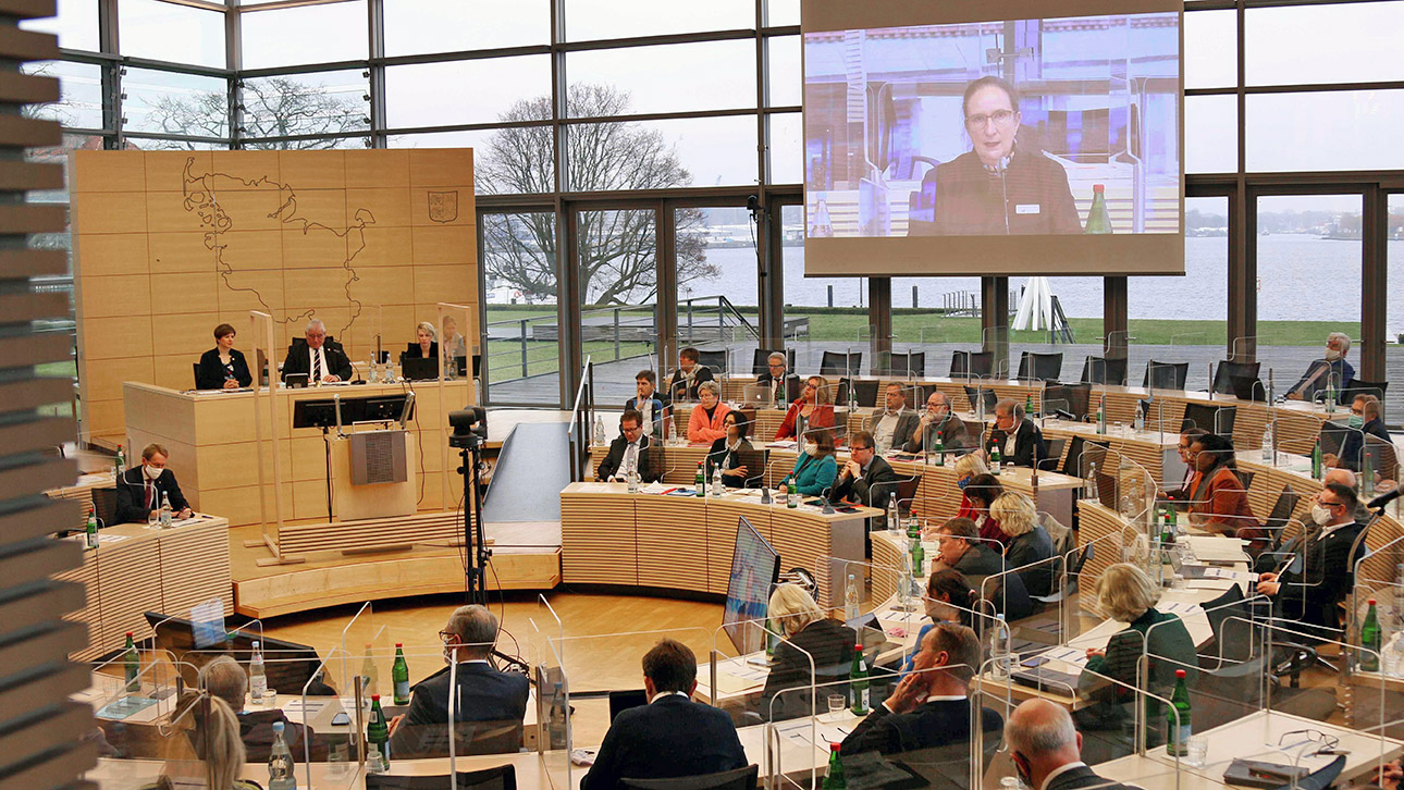 Blick in den Plenarsaal, wo Abgeordnete im Kreisrund sitzen und auf einer großen Leinwand die Corona-Expertin Alexandra Barth zugeschaltet ist.