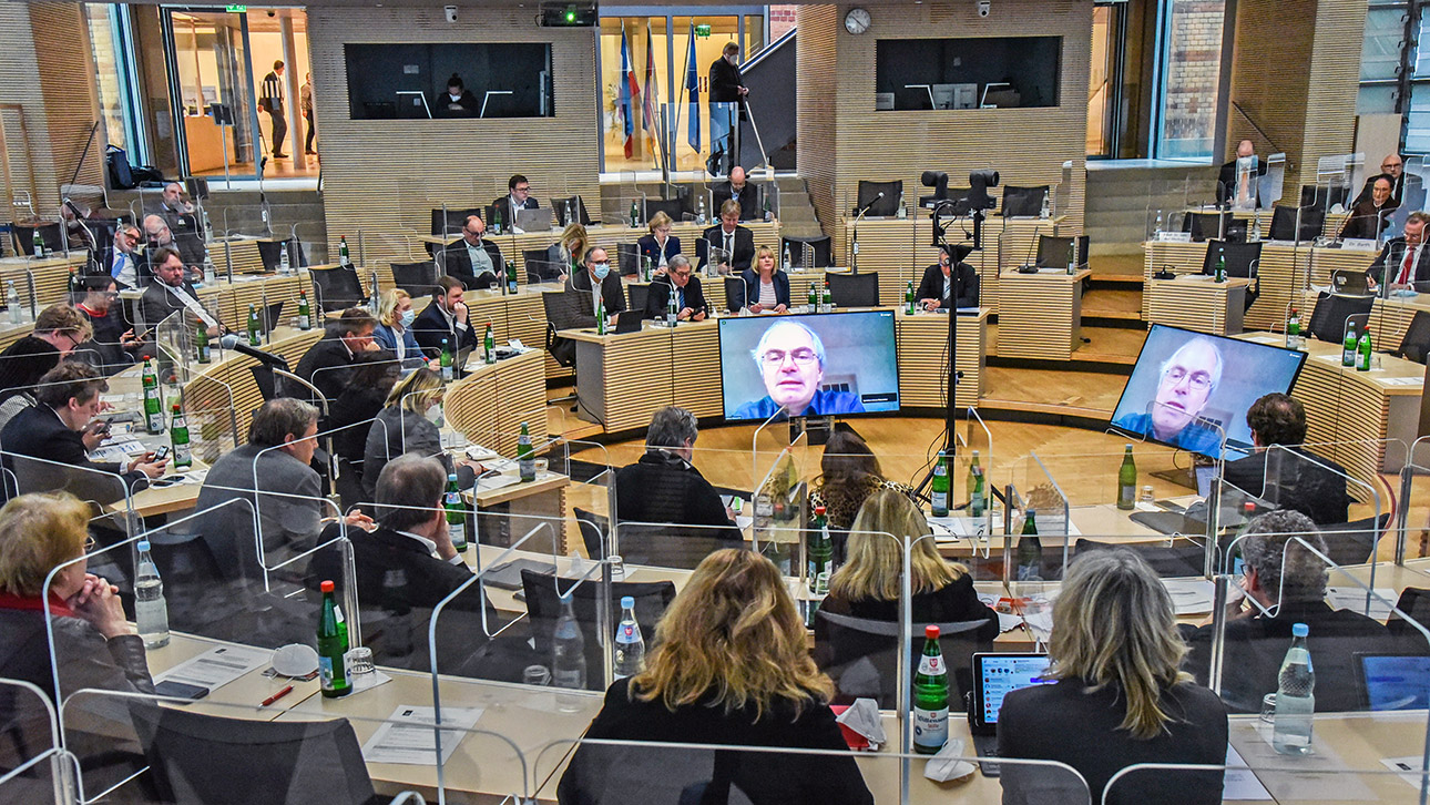 Die Abgeordneten sitzen im Plenarsaal des Schleswig-Holsteinischen Landtages in einem Rund zusammen. In der Mitte stehen zwei Monitore, auf dem Helmut Fickenscher, Leiter des Instituts für Infektionsmedizin an der Kieler Uni. zu sehen ist.