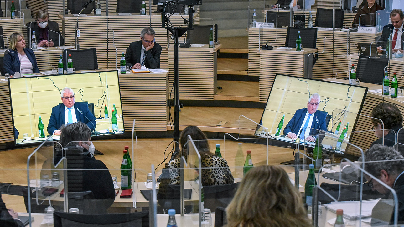 Landtagspräsident Klaus Schlie ist auf zwei Bildschirmebn im Plenarsaal des Landtages zu sehen. Um ihn herum sitzen Abgeordnete im Rund.