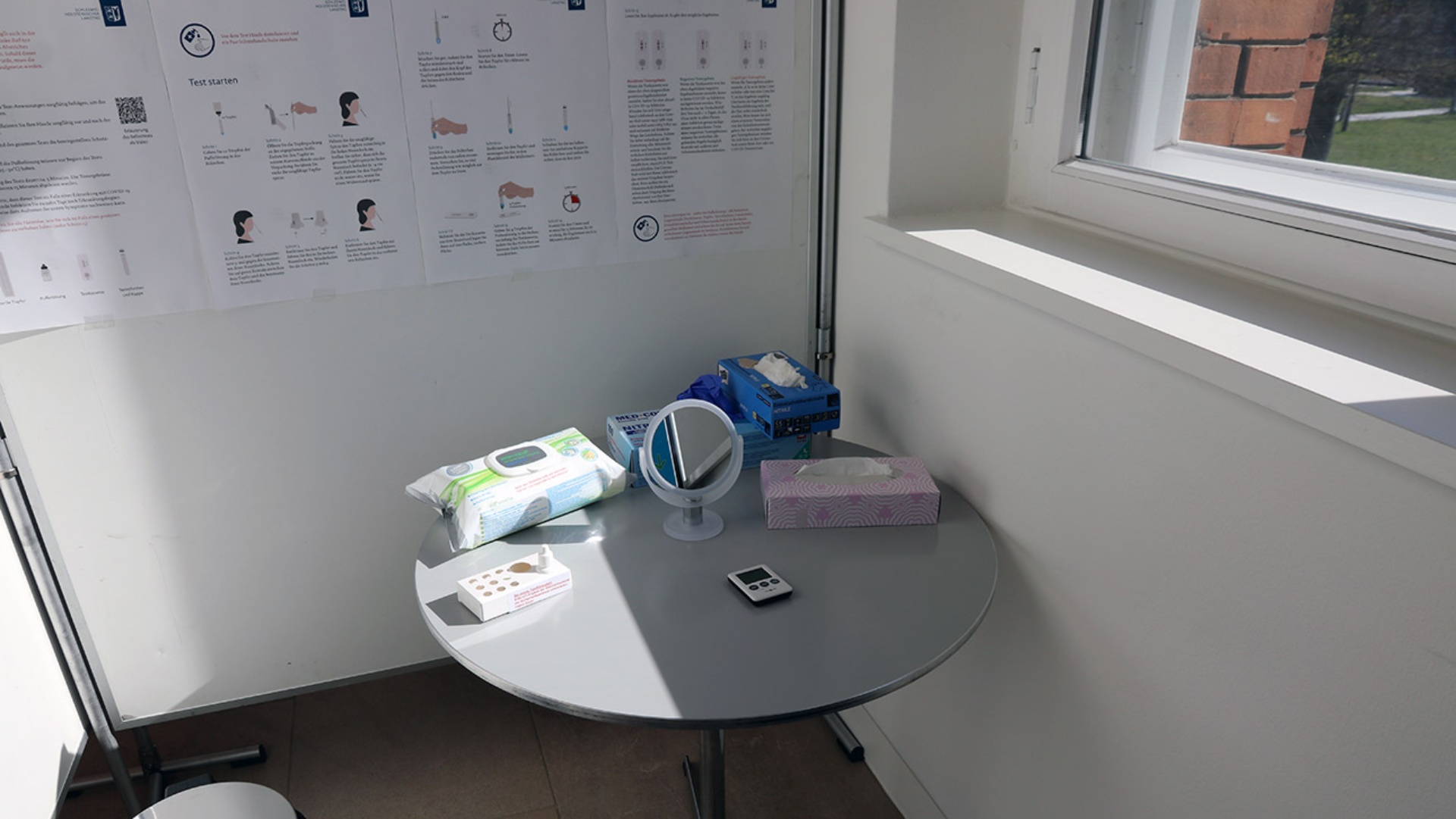 Blick in eine Corona-Test-Kabine im Landtag, wo auf einem Tisch der Test, ein Spiegel und Tücher bereitliegen. An der Wand hängt eine Anleitung im Großformat.stenlos testen können.