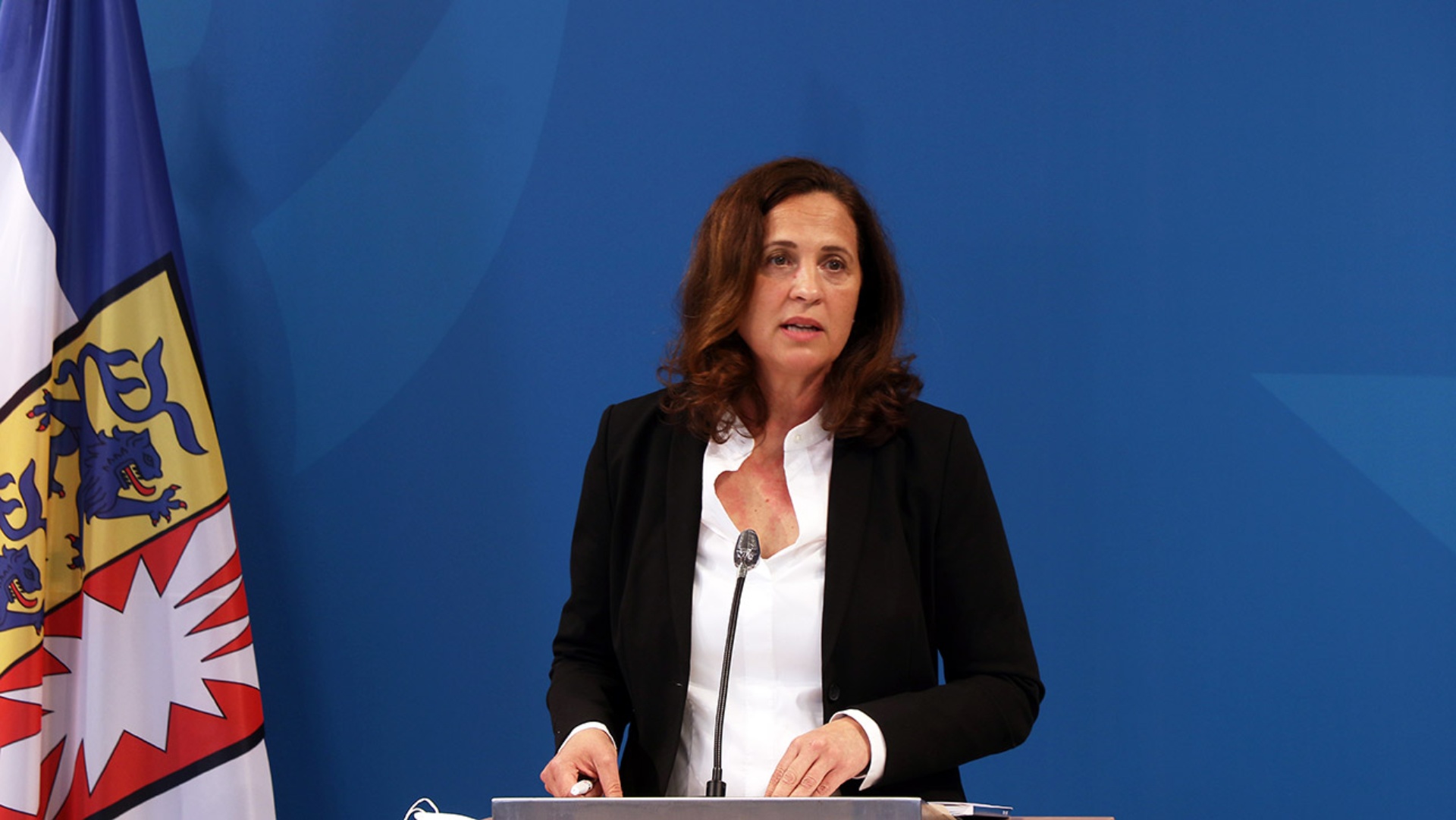 Die Polizeibeauftragte steht vor einer blauen Leinwand an einem Podium und stellt ihren Tätigkeitsbericht vor. Links im Bild die Schleswig-Holstein-Flagge.