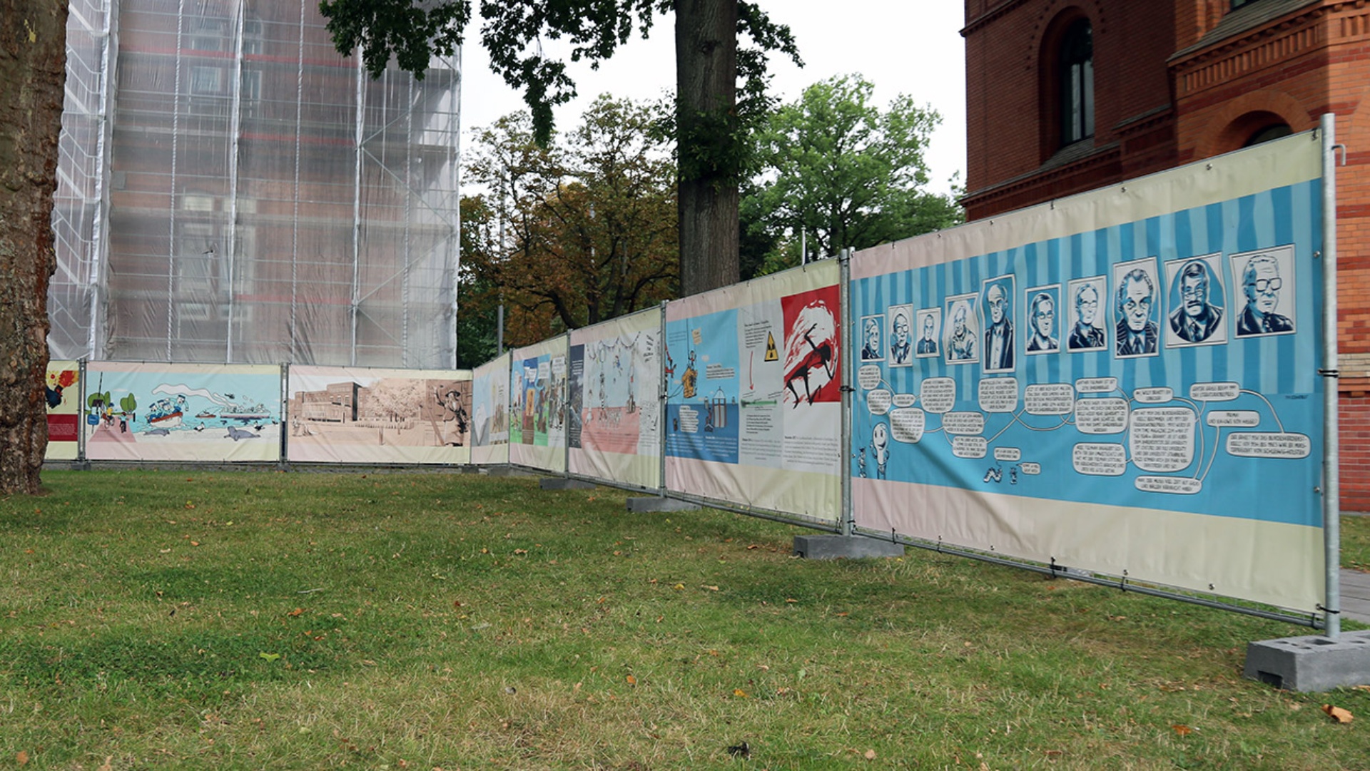 Auf einenm Bauzaun vor dem mit einem Gerüst versehenen Landtagsgebäude hängen Comics, die zum 75. Landesjubiläum herausgegeben wurden, an einem Bauzaun.