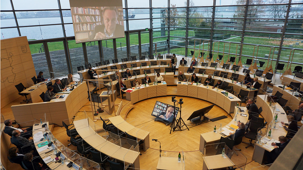 Die Anhörung im Plenarsaal des Landtages wird per Hybrid veranstaltet. An der Decke und im Plenarrund sind Bildschirme aufgebaut..