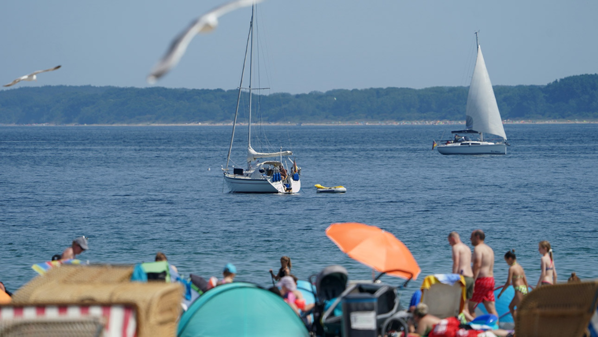 Urlauber und Tagestouristen genießen das Sommerwetter am Ostseestrand in Travemünde. Auf dem Wasser dümpeln Segelboote, in der Luft fliegen Möwen.