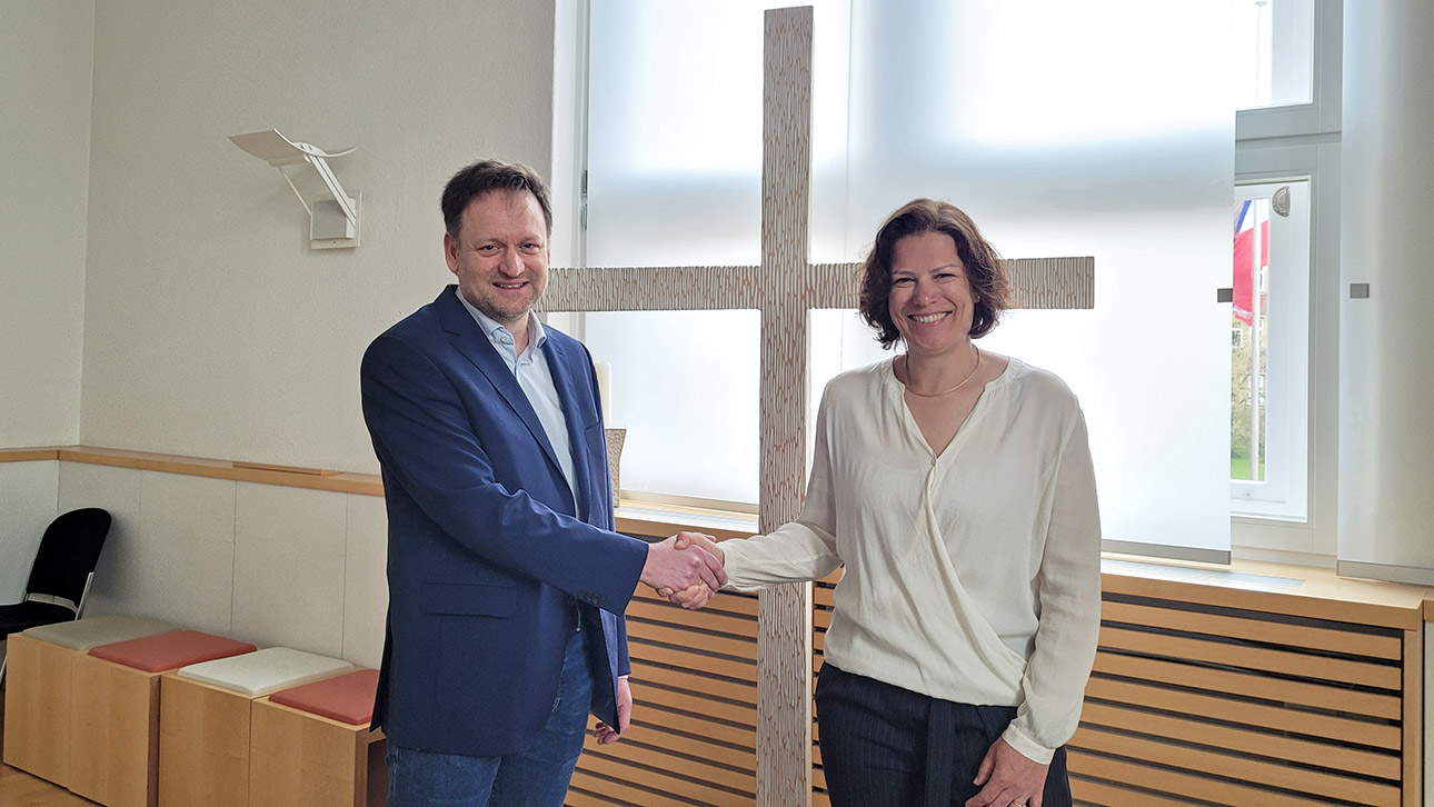 Landtagspräsidentin Kristina Herbst begrüßt Pastor Wilko Teifke im Landeshaus. Sie stehen vor einem Kreuz.