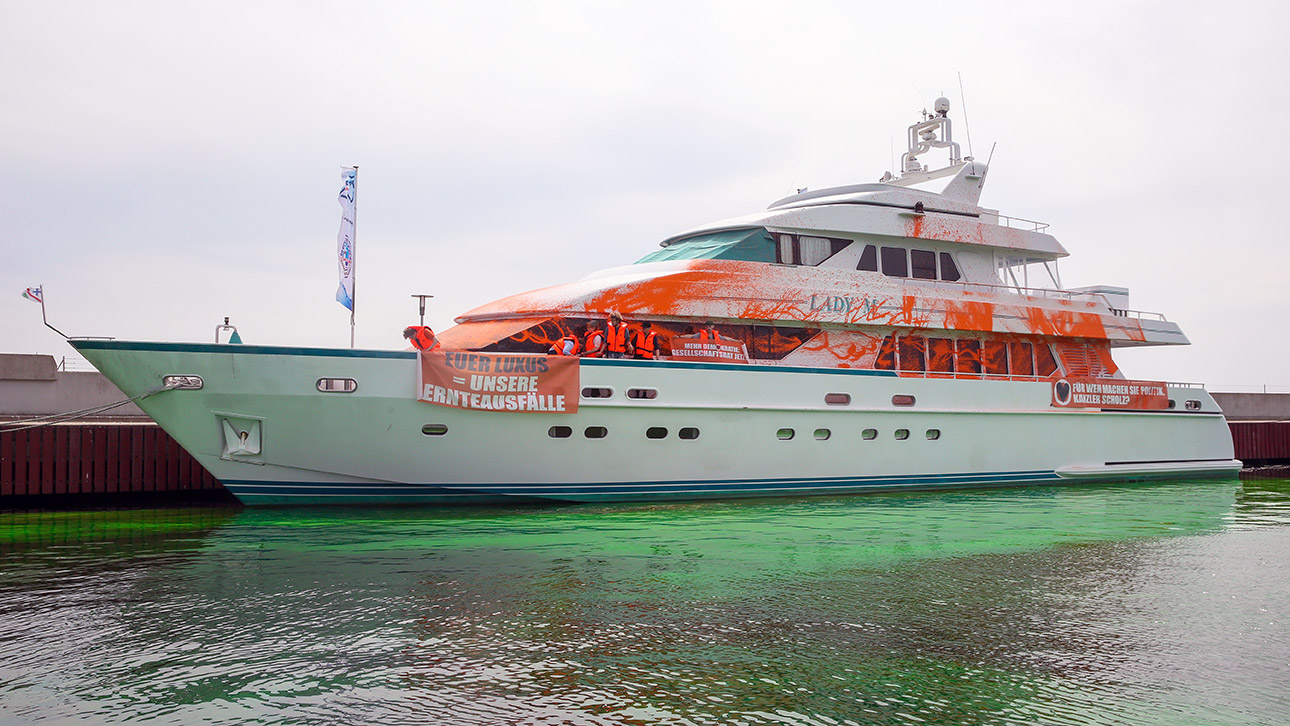 Teilnehmer einer Protestaktion stehen auf einer von ihnen mit oranger Farbe besprühten Yacht im Hafen von Neustadt. Auf einem Transparent ist die Aufschrift „Euer Luxus = Unsere Ernteausfälle“ zu sehen. 