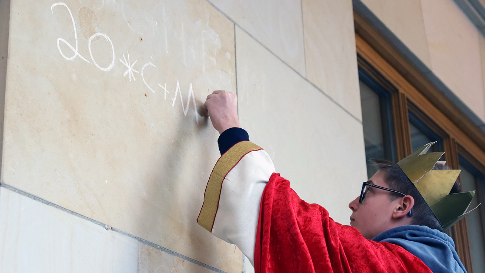 Ein Sternsinger schreibt mit weißer Kreide den Segen „20*C+M+B+24“ an den Haupteingang des Landeshauses.