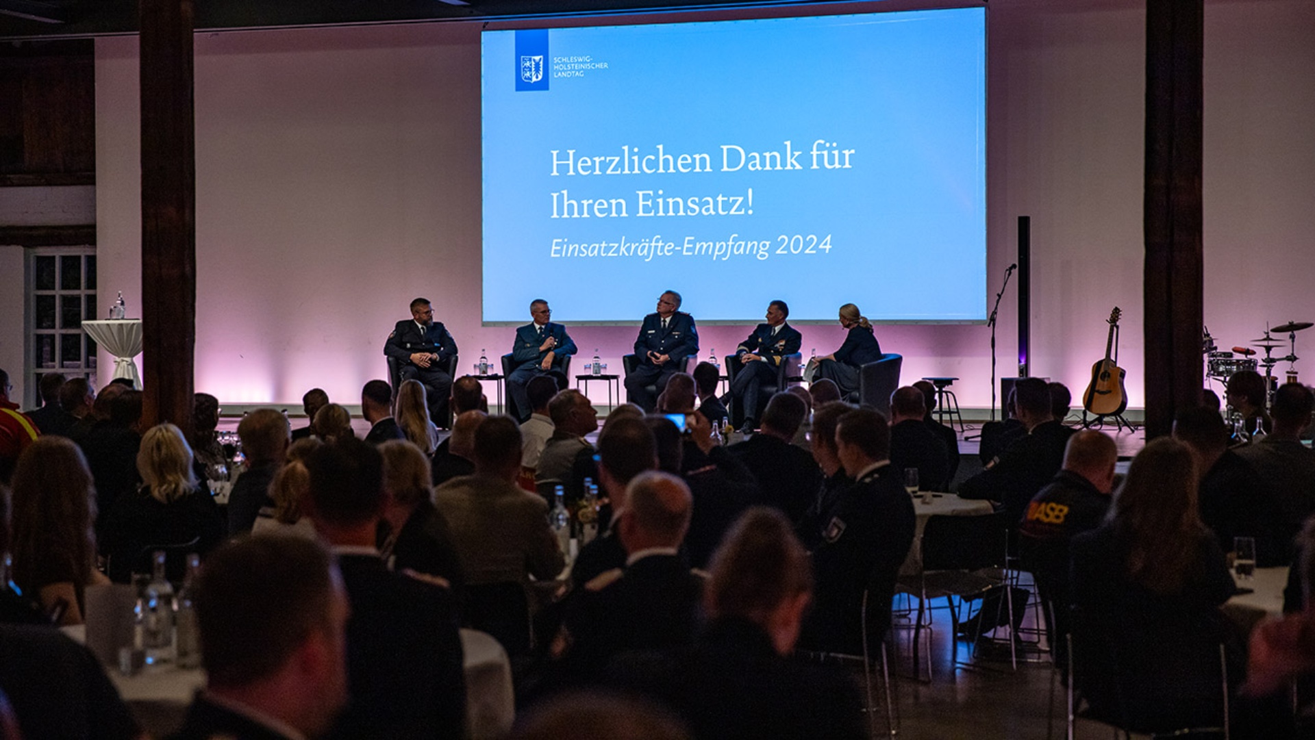 Gut 250 Vertreterinnen und Vertreter von Bundeswehr, Polizei, Bundesfreiwilligendienst, Katastrophenschutzeinheiten und Zoll verfolgen eine Podiumsdiskussion. Über der Bühne ist auf eine Leinwand projeziert: "Herzlichen Dank für Ihren Einsatz"  