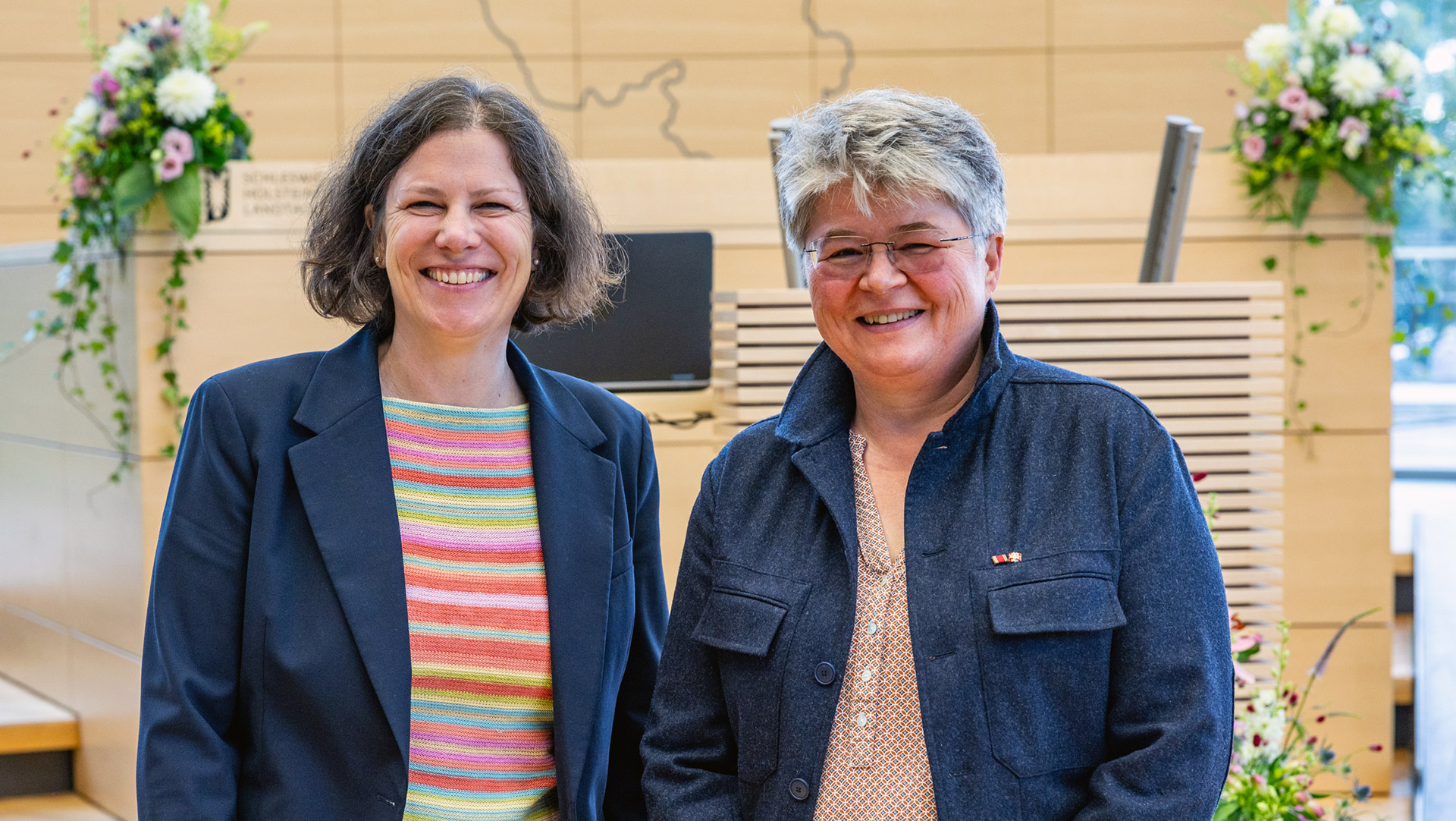 Landtagspräsidentin Kristina Herbst und Referentin Friederike Sittler stehen im Plenarsaal vor dem Rede-Pult und schauen in die Kamera.