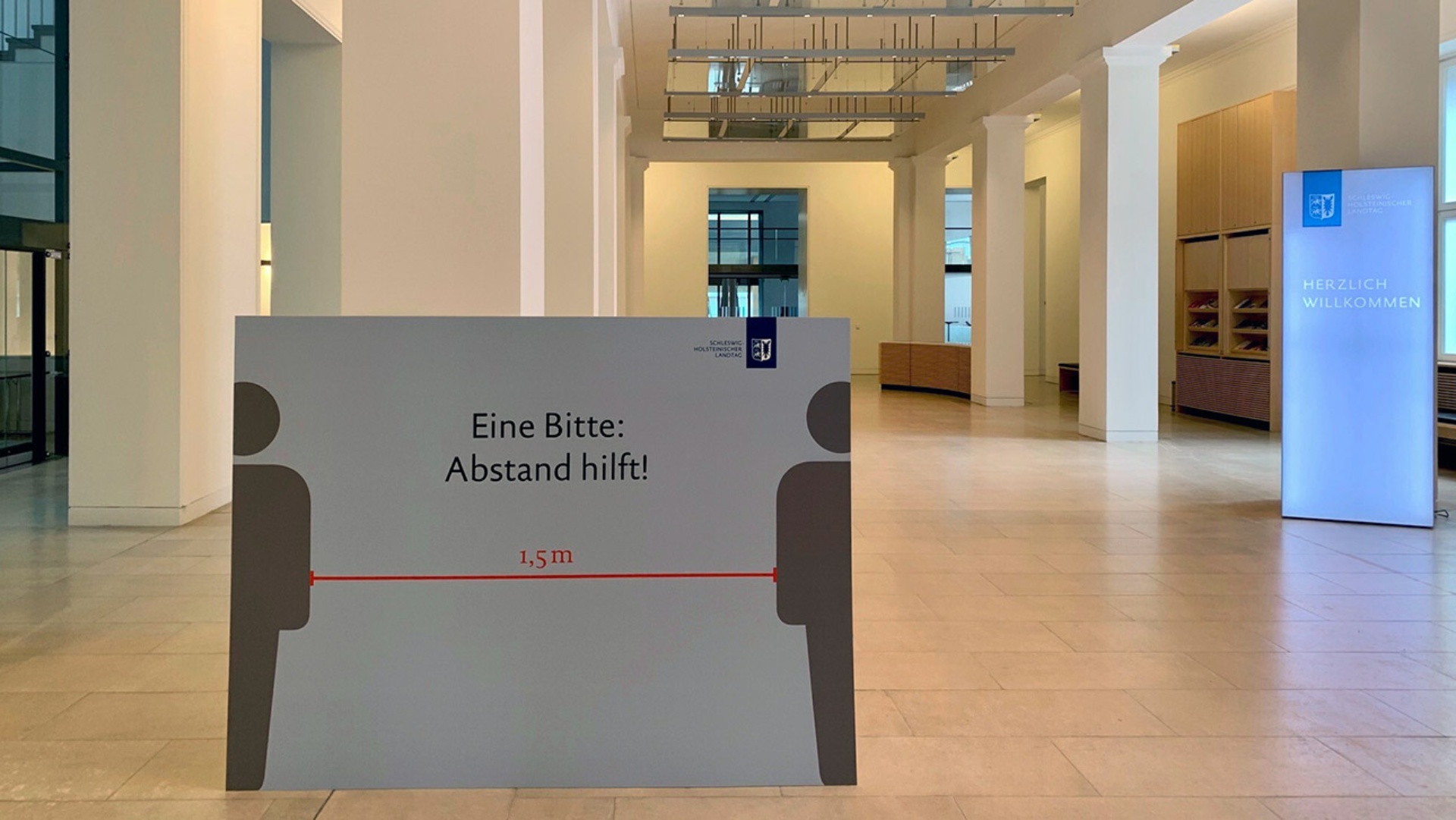 Ein Schild mit der Aufschrift  "Eine Bitte: Abstand hilft" steht in der Eingangshalle des Landeshauses in Kiel.