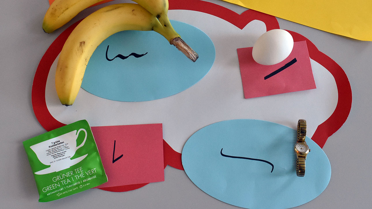 Eine Banane, ein Ei, eine Uhr und ein Teebeutel liegen auf buntem Papier. Darauf geschrieben stehen die Bezeichnungen in Stenografie