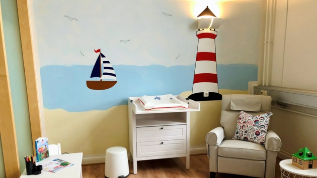 In einem Pastellfarben gestrichenen Raum sind ein Spieltisch, eine Wickelkommode und ein großer Sessel zu sehen. An die Wandgemalt ist ein Leuchtturm mit einer echten Lampe an der Spitze..